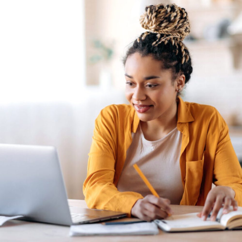 Focused cute stylish african american female student with afro dreadlocks, studying remotely from home, using a laptop, taking notes on notepad during online lesson, e-learning concept, smiling